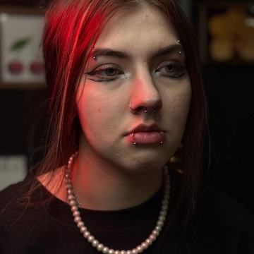 Close-up portrait in red studio lighting of a person with multiple facial piercings—eyebrow bars, a septum ring, and two lower-lip studs—illustrating professional piercing options at VeAn in Vilnius.
