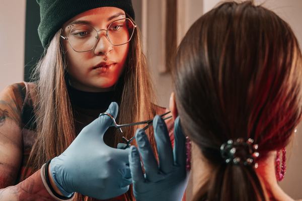 Gloved piercer in glasses positions forceps on a client’s ear under warm studio lighting, illustrating sterile, professional ear‑piercing services in Panevėžys.