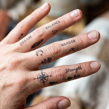 Mažos juodo rašalo tatuiruotės ant pirštų ir plaštakos: žodžiai DREAM, BALANCE, WANDER, užrašas „Strength“, kompasas, mėnulis, banga ir trikampis—minimalistinio blackwork stiliaus pavyzdys matomoje vietoje.