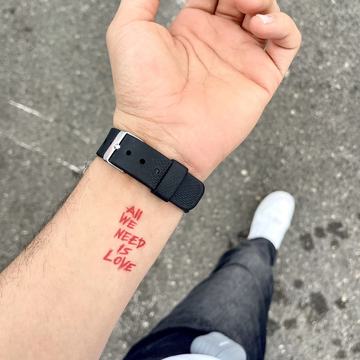 Maža raudono rašalo tekstinė tatuiruotė ant vidinės dilbio pusės prie riešo: „ALL WE NEED IS LOVE“ keturiomis eilutėmis didžiosiomis raidėmis, minimalistinis lettering stilius; šalia matomas juodas laikrodžio dirželis.
