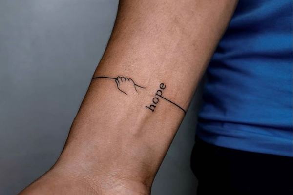 Maža juodo rašalo tatuiruotė ant vidinės dilbio pusės prie riešo: plona linija su mažos rankos kontūru, pereinanti į vertikalų užrašą „hope“—minimalistinio blackwork stiliaus pavyzdys matomoje vietoje.