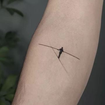 Maža juodo rašalo tatuiruotė ant vidinės dilbio pusės: minimalistinis lyno akrobato siluetas su ilgu pusiausvyros strypu—blackwork pavyzdys mažam formatui, simbolizuojantis balansą.