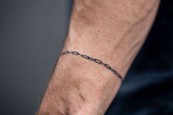 Maža juodo rašalo grandinėlės tatuiruotė aplink riešą – minimalistinio blackwork stiliaus pavyzdys matomoje dilbio zonoje.