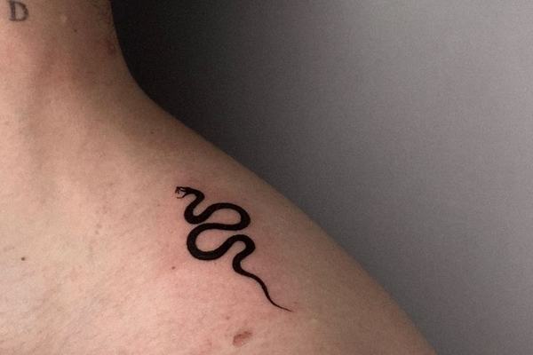 Maža juodo rašalo gyvatės kontūro tatuiruotė ant peties prie raktikaulio—minimalistinis blackwork pavyzdys matomoje vietoje.