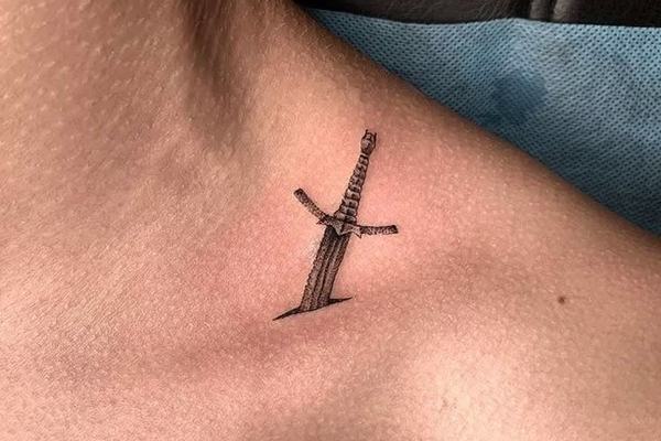 Maža juodo rašalo kardo tatuiruotė ant kaklo prie raktikaulio – minimalistinio blackwork stiliaus pavyzdys matomoje vietoje.