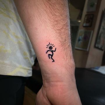 Maža juodo rašalo tatuiruotė ant vidinės dilbio pusės: šokanti lazdelinė figūra su spindulių linijomis virš galvos—minimalistinio blackwork stiliaus pavyzdys mažam formatui matomoje vietoje.