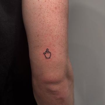 Maža juodo rašalo tatuiruotė ant žasto nugarinės pusės: pikselinio stiliaus rankos ikona su iškeltu viduriniu pirštu—minimalistinio blackwork mikrotatuiruotės pavyzdys aiškiais kontūrais.