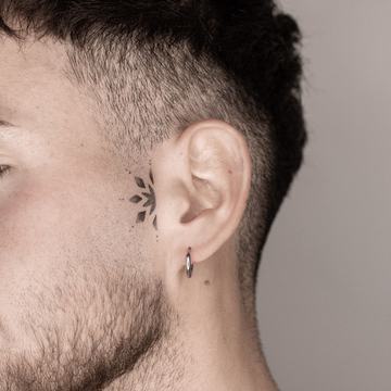 Maža juodo rašalo tatuiruotė prie ausies: grafiškas lapelių motyvas ties smilkiniu—minimalistinio blackwork stiliaus pavyzdys matomoje vietoje.