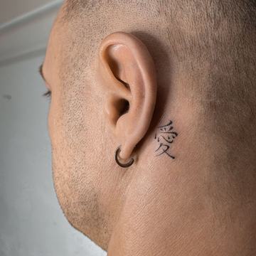 Maža juodo rašalo tatuiruotė už ausies: vertikaliai ištatuiruotas kanji ženklas—minimalistinio blackwork stiliaus pavyzdys mažam formatui matomoje vietoje.