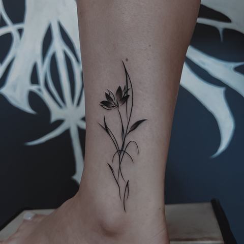 Subtili augalinė tatuiruotė ant blauzdos su plonu stiebu, smulkiais lapeliais ir lengva, natūralia kompozicija.