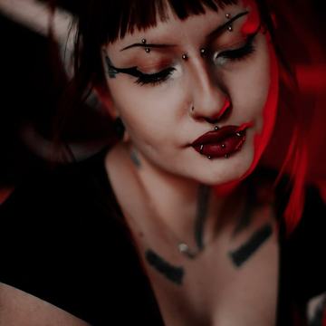 Artimas moters portretas raudonai apšviestoje piercingo studijoje: septum žiedas, keli apatinės lūpos (labreto) auskarai, antakių ir nosies auskarai, ryškus akių makiažas ir tatuiruotės ant krūtinės.