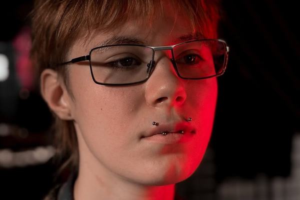 Close-up of a person in glasses with two lower‑lip piercings (snake bites) under red studio lighting, illustrating facial piercing options at a professional studio in Panevėžys.