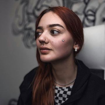 Close-up studio portrait of a person with septum and nostril rings, a cheek dermal, and earlobe studs, illustrating professional facial piercing options at VeAn in Panevėžys.