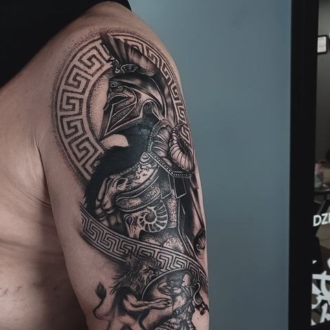 Detali juodai pilka tatuiruotė ant žasto su kario motyvu, geometriniais ornamentais ir giliu šešėliavimu, pabrėžiančiu kompozicijos charakterį.