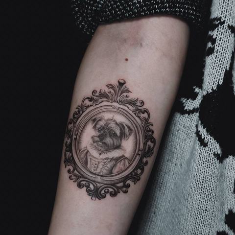 Apvali simbolinė tatuiruotė ant dilbio išsiskiria kruopščiai išpieštomis detalėmis, subtiliu šešėliavimu ir dekoratyviu rėmeliu, suteikiančiu motyvui užbaigtumo.