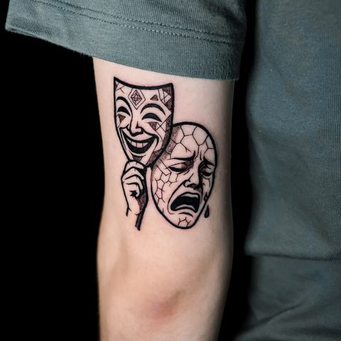 A blackwork upper arm tattoo featuring contrasting theatrical masks with expressive details