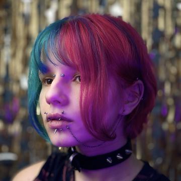 Close-up portrait in purple lighting of a person with split-dyed teal and magenta hair, a spiked choker, and multiple facial and ear piercings—eyebrow/bridge studs, several lip and labret piercings with spikes and a chain, and helix/lobe studs—illustrating professional piercing options at VeAn in Vilnius.