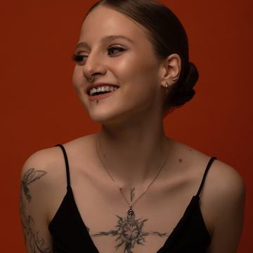 Artimas besišypsančios moters portretas su septum žiedu, labreto (apatinės lūpos) auskaru, keliomis ausų auskarais ir tatuiruotėmis ant krūtinės bei peties, oranžiniame studijos fone.