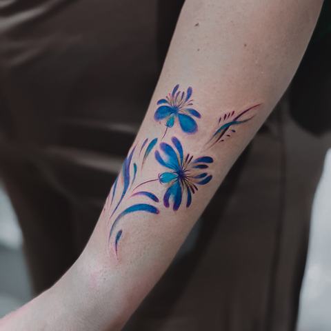A vibrant floral forearm tattoo with flowing blue and purple leaves and delicate linework