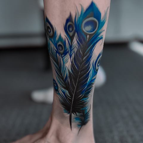 Ryški povo plunksnų tatuiruotė ant blauzdos, išsiskirianti sodriais mėlynais ir žalsvais atspalviais, grakščia forma ir elegantiškai išdėstyta kompozicija.