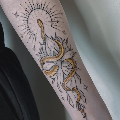 An elegant fine line forearm tattoo featuring a radiant figure with flowing ornamental details and subtle golden tones