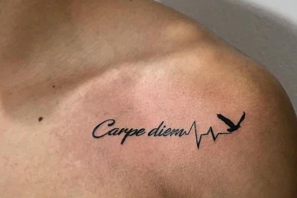 Maža juodo rašalo tatuiruotė ant krūtinės prie raktikaulio: kursyvu parašyta frazė „Carpe diem“, pereinanti į širdies ritmo liniją ir paukščio siluetą—minimalistinio blackwork stiliaus pavyzdys mažam formatui matomoje vietoje.